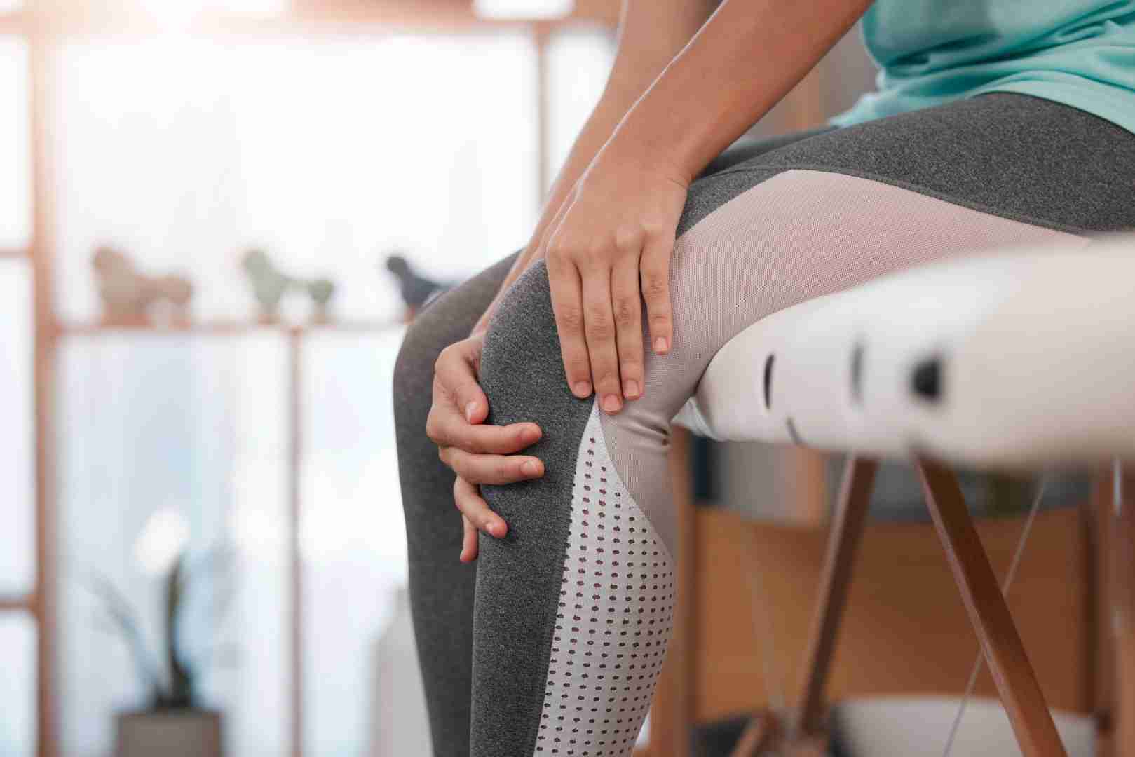 A person wearing grey and white leggings is seated, holding their knee with both hands. The indoor setting is softly lit, with sunlight streaming through a window in the background. The scene suggests a moment of rest or addressing knee discomfort, much like a session at Austin Manual Therapy Associates.