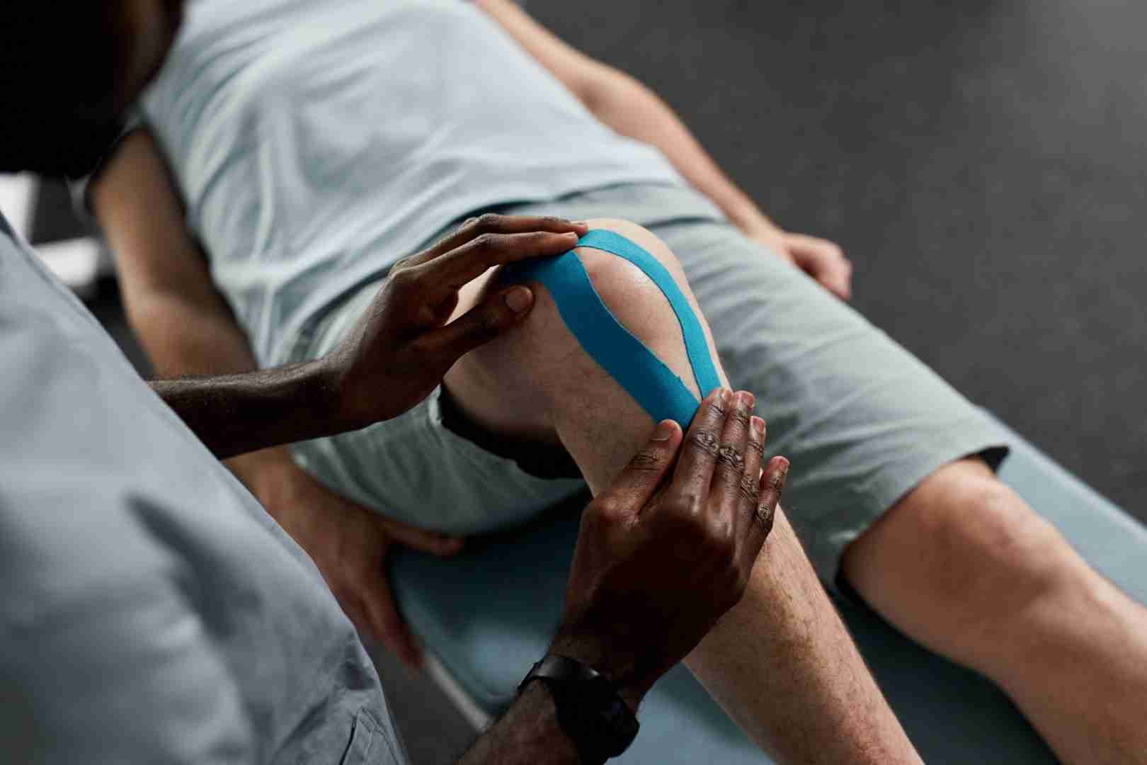 A person is lying on a treatment table at Austin Manual Therapy Associates while another individual applies blue kinesiology tape to their knee. The person receiving the treatment is wearing light shorts and a matching shirt, in what appears to be a clinical or therapeutic setting.