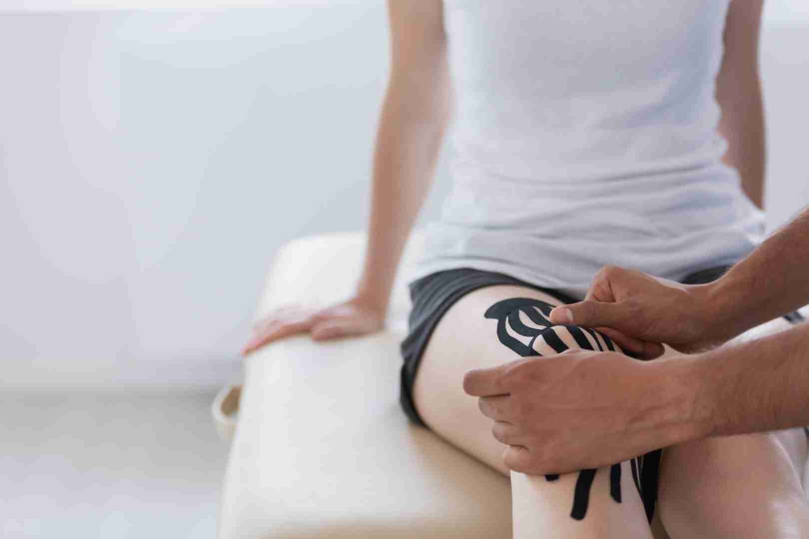 A person sitting on an examination table at Austin Manual Therapy Associates is being treated with kinesiology tape applied to their knee by another person. The individual receiving the treatment is wearing a white tank top and black shorts.