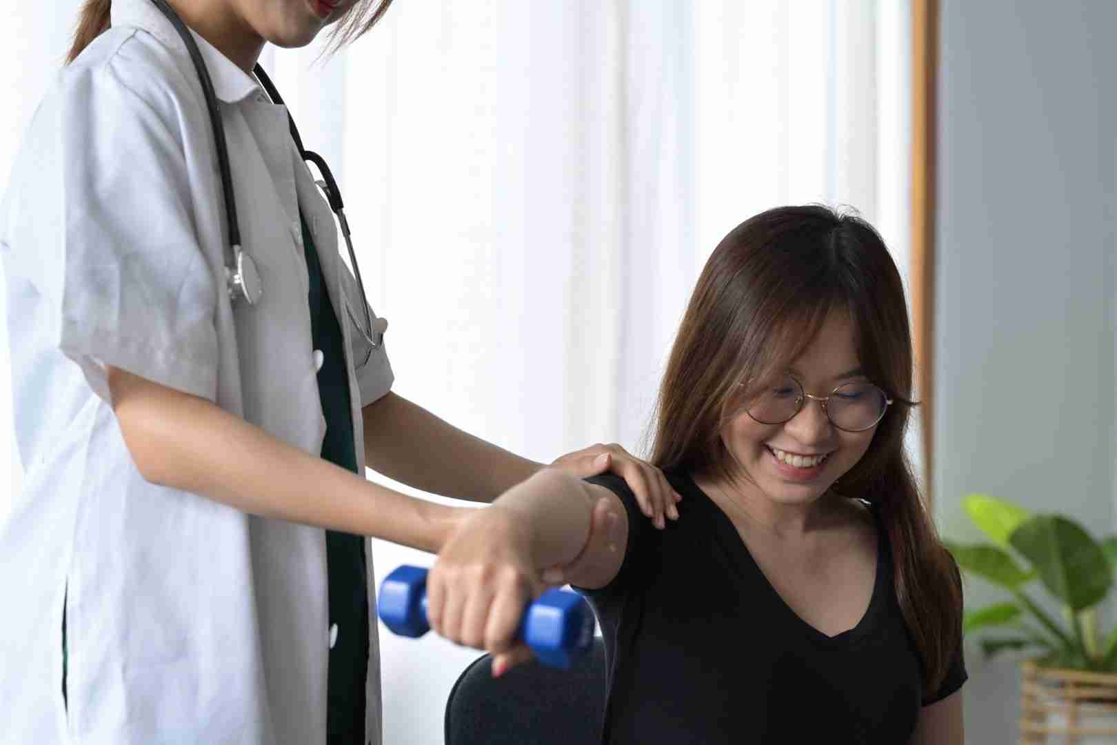 A healthcare professional from Austin Manual Therapy Associates assists a smiling woman with physical therapy exercises. The woman, wearing glasses and a black shirt, holds a blue dumbbell while the professional guides her arm. The setting appears to be a medical or rehabilitation clinic.