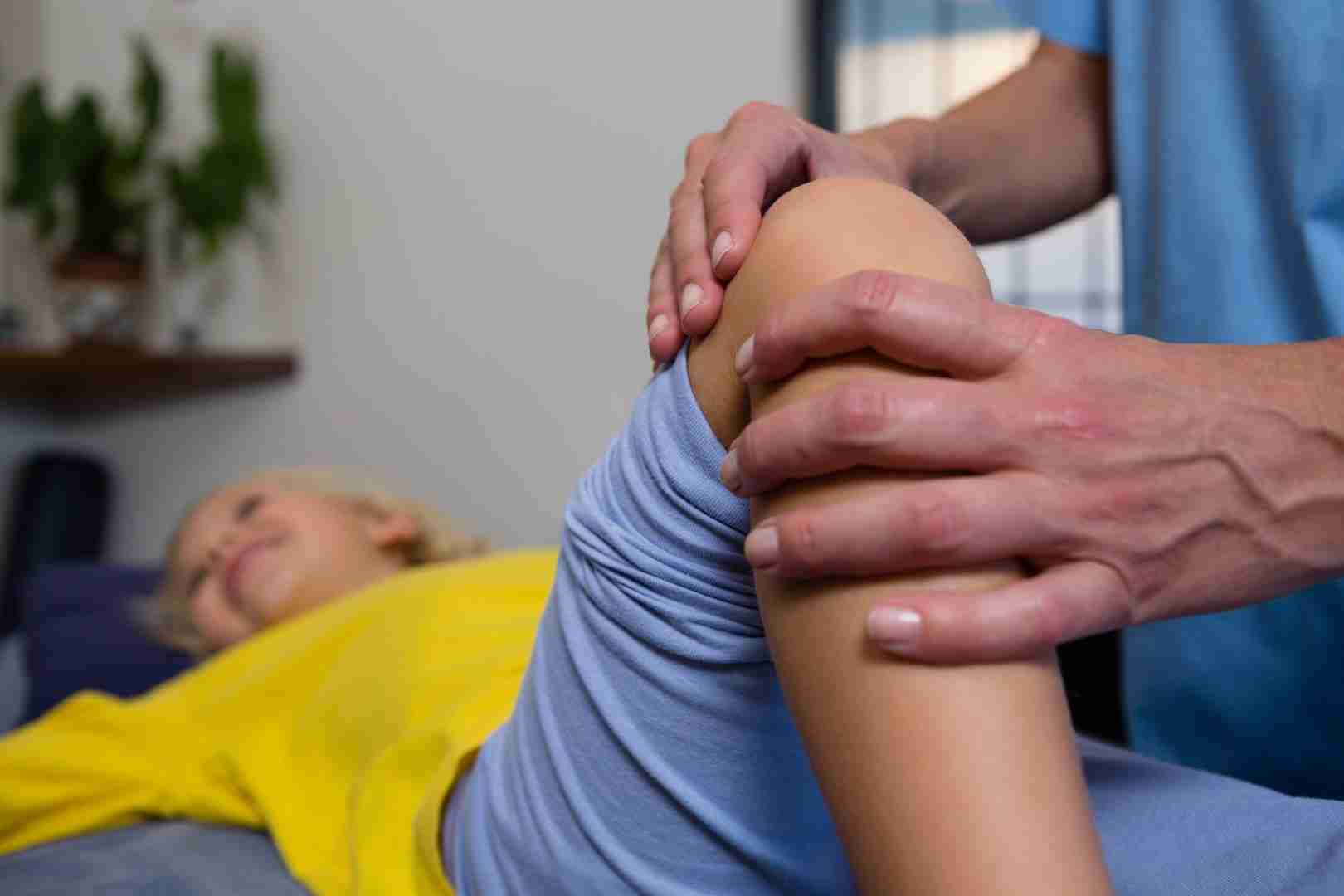A physical therapist from Austin Manual Therapy Associates in blue scrubs uses their hands to examine the knee of a patient wearing a yellow shirt. The patient is lying on a treatment table with their leg bent at the knee and appears to be relaxed.